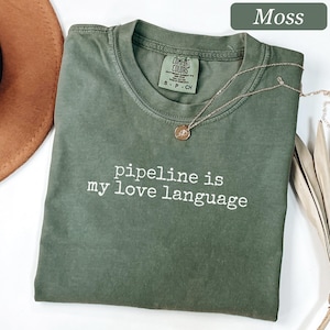 May include: A moss green short-sleeve t-shirt with a crew neckline and the white text "pipeline is my love language" printed on the chest. The shirt features a Comfort Colors brand label inside the collar and a thin gold-toned chain necklace.