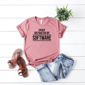 May include: A dusty rose t-shirt with the text "EASILY DISTRACTED BY SOFTWARE" in black. The shirt is paired with blue jeans, brown sandals, and a small potted plant on a white wooden surface.