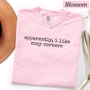 May include: A light pink short-sleeved t-shirt with a crew neckline. The shirt features the black text "apparently, i like cozy corners" printed across the chest. The brand label "Comfort Colors" is visible on the inside of the collar.