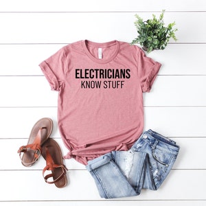 May include: A dusty rose t-shirt with the text "Electricians Know Stuff" printed in black.