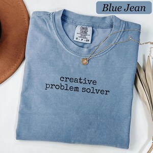 May include: A blue jean colored t-shirt with a crew neckline and short sleeves. The shirt features the black embroidered text "creative problem solver" on the chest. A Comfort Colors brand label is visible at the inner neck, indicating size S.