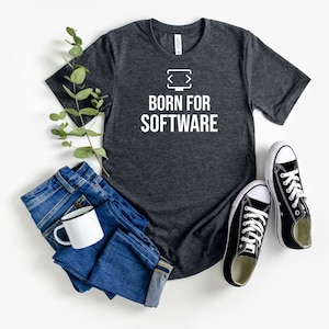 May include: A dark heather grey t-shirt with a white graphic of a computer screen with angle brackets and the text "Born for Software". The shirt is laid flat on a white surface with a pair of black sneakers and a pair of blue jeans.