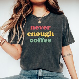 May include: A dark gray t-shirt with the text "never enough coffee" in colorful block letters. The text is in a retro style font.