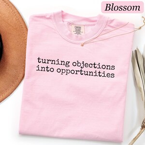 May include: A light pink t-shirt with a crew neckline and short sleeves. The shirt features the black text "turning objections into opportunities" printed in a typewriter-style font across the chest. A Comfort Colors brand label is visible.
