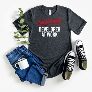 May include: A dark heather grey t-shirt with a white graphic that says "WARNING DEVELOPER AT WORK" in red letters.