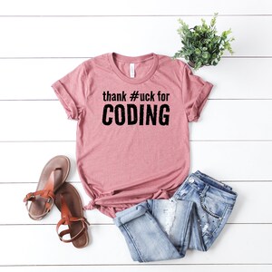 以下が含まれることがあります： ピンクのTシャツに黒い文字で「thank #uck for CODING」と書かれています。