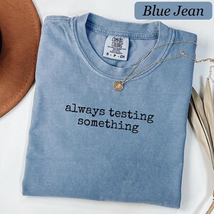 Peut inclure: Un t-shirt à manches courtes de couleur bleu jean avec une encolure ronde. Le vêtement présente le texte brodé en noir "always testing something" centré sur la poitrine. Un collier doré avec un petit pendentif circulaire repose sur le col.