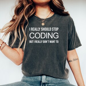 以下が含まれることがあります： 「I REALLY SHOULD STOP CODING BUT I REALLY DON'T WANT TO」の白文字がプリントされた、濃いグレーのTシャツ。クルーネックのスタイルで、人が着用しています。