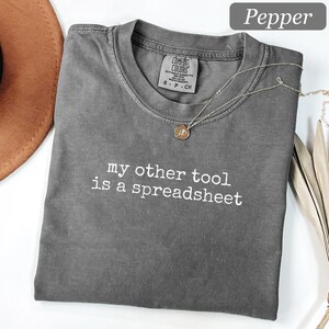 以下が含まれることがあります： チャコールグレーの半袖Tシャツ。クルーネックで、胸の中央に白い刺繍で「my other tool is a spreadsheet」という文字が入っています。襟の内側にはComfort Colorsのブランドラベルがあり、細いゴールドのネックレスが添えられています。