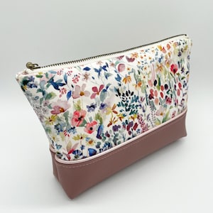 May include: A floral print cosmetic bag with a mauve-coloured bottom and a bronze zip. The bag features a white background with colourful flowers in shades of pink, blue, and yellow. The bag is designed to hold makeup and other beauty products.