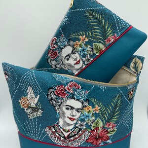 May include: Two teal blue zippered pouches with a floral and portrait design of Frida Kahlo. The larger pouch has a red trim and the smaller pouch has a grey trim.