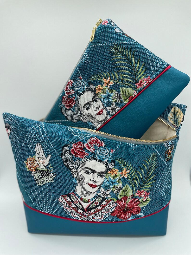 May include: Two teal blue zippered pouches with a floral and portrait design of Frida Kahlo. The larger pouch has a red trim and the smaller pouch has a grey trim.