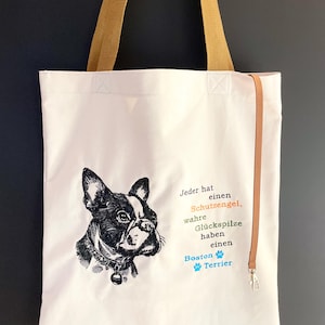 May include: Cream tote bag with a black and white Boston Terrier illustration. It has tan handles and a brown strap with a silver clasp. The German text reads "Jeder hat einen Schutzengel, wahre Glückspilze haben einen Boston Terrier."