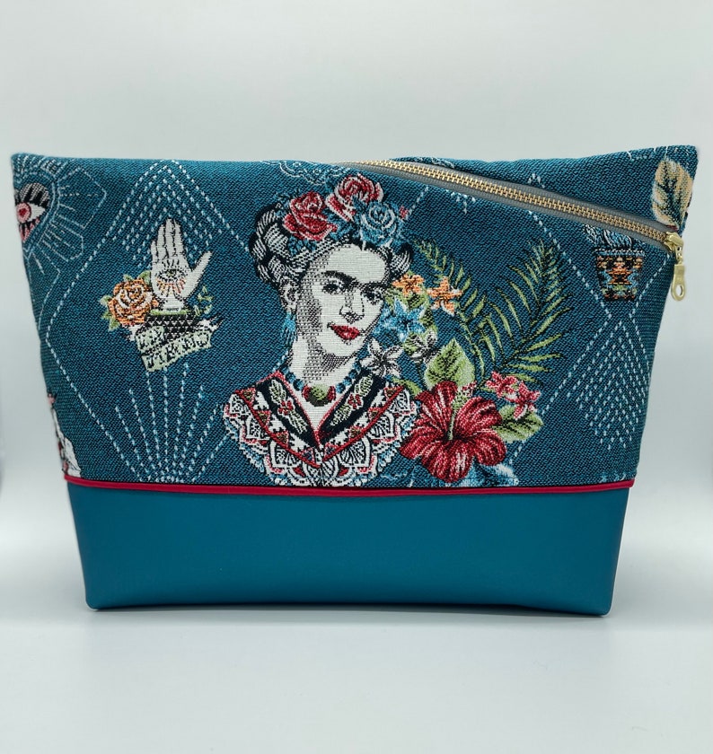 May include: A teal blue zippered pouch with a colourful tapestry design featuring a portrait of Frida Kahlo. The pouch has a red trim and a teal blue bottom.