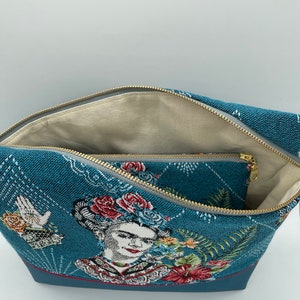 May include: A blue zippered pouch with a woven design featuring a portrait of Frida Kahlo with flowers. The pouch has a smaller zippered pocket inside.
