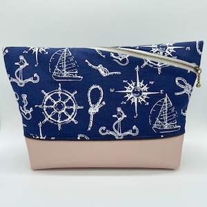 May include: A blue zippered pouch with a pink base. The pouch is decorated with a white nautical pattern of anchors, ships, wheels, and compass roses.