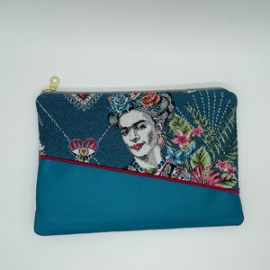 May include: A blue and white tapestry clutch bag with a zip closure. The tapestry features a portrait of Frida Kahlo with flowers and a heart design. The bag has a blue leather base with a red stitch detail.
