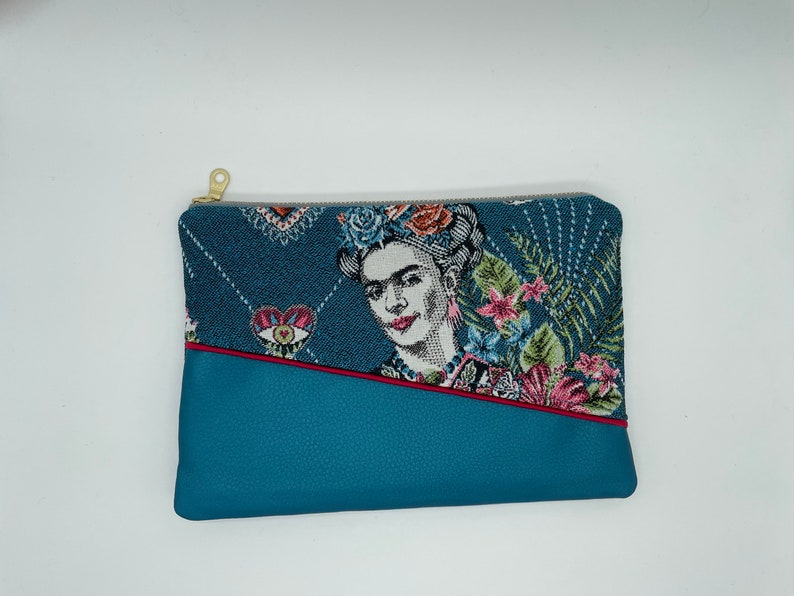 May include: A blue and white tapestry clutch bag with a zip closure. The tapestry features a portrait of Frida Kahlo with flowers and a heart design. The bag has a blue leather base with a red stitch detail.
