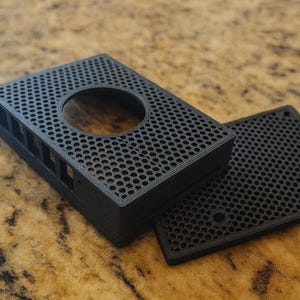 May include: Black rectangular electronic device enclosure with a perforated design and a circular opening. A separate, matching perforated cover is next to it. The device is likely for electronics or a small computer.