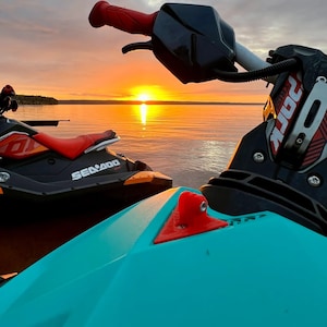 May include: Two jet skis on a lake at sunset. The jet ski in the foreground is turquoise blue with a red handle. The jet ski in the background is black with orange accents and the text "Sea-Doo" printed on the side.