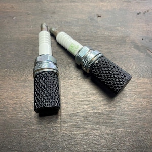 May include: Two black spark plugs with a diamond pattern on the metal casing. The spark plugs are on a wooden surface.