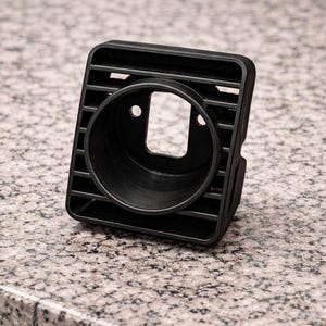Puede incluir: Una rejilla de ventilación de coche de plástico negro con una abertura circular y un marco rectangular. La rejilla tiene lamas horizontales y está diseñada para un salpicadero. Se encuentra sobre una superficie moteada.