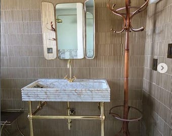 May include: A white marble bathroom sink with a gold metal stand and a gold faucet. The sink is in a bathroom with brown tile walls and a wooden coat rack.