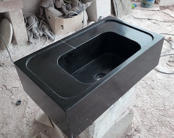 May include: A black rectangular stone sink with a rounded edge and a single drain hole. The sink is sitting on a white stone base.