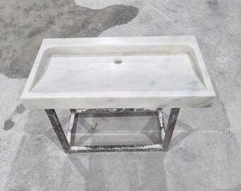 Può includere: Un lavabo rettangolare in marmo bianco con un foro di scarico, posto su un telaio in metallo scuro. Il lavabo ha una superficie liscia con venature sottili. Il telaio in metallo ha una base rettangolare.