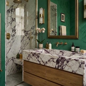 May include: A bathroom vanity featuring a white marble countertop with deep purple veining, integrated sink, and light wood drawers. The walls are painted a deep green, complemented by a gold-framed mirror, brass fixtures, and wall sconces.