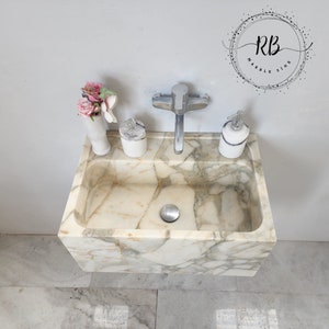 May include: A rectangular marble sink with a cream and gray veined pattern. The sink has a chrome faucet and is accessorized with a white vase of pink flowers, a soap dispenser, and a small container. The logo "RB Marble Sink" is visible.