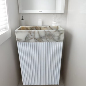 May include: A white pedestal sink with a rectangular basin. The upper portion features a marble pattern in shades of white, gray, and brown. The lower section has a vertical ribbed design. The sink is set against a white tiled wall.
