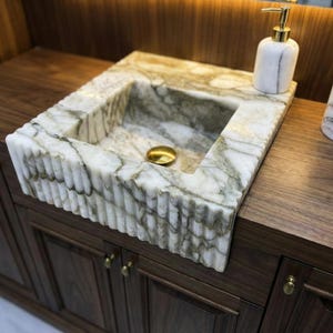 Può includere: Un lavabo rettangolare in marmo con scarico dorato. Il lavabo è bianco con venature grigie e verdi e poggia su un mobile in legno scuro. Un dosatore di sapone e un contenitore abbinati sono sul piano di lavoro.