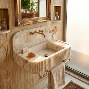 May include: A marble bathroom sink with a gold faucet and towel rack. The sink has a rectangular basin and a ribbed front. A bar of soap and a brush sit on the sink's ledge. A beige towel hangs from the rack.