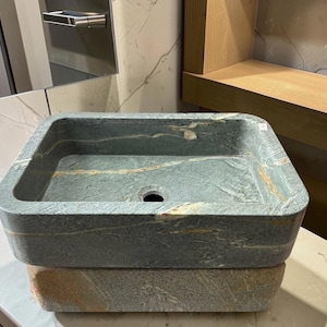 Peut inclure: Un &eacute;vier en pierre de forme carr&eacute;e, avec des nuances de gris et d'or. Il pr&eacute;sente un trou d'&eacute;vacuation central et repose sur une base en pierre assortie. Le design met en valeur les veines naturelles, pour un style rustique et &eacute;l&eacute;gant.