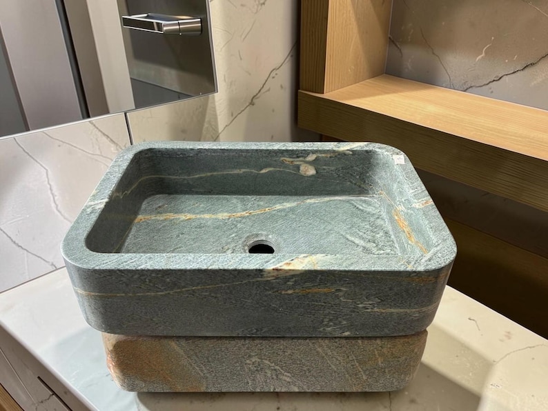 Peut inclure: Un &eacute;vier en pierre de forme carr&eacute;e, avec des nuances de gris et d'or. Il pr&eacute;sente un trou d'&eacute;vacuation central et repose sur une base en pierre assortie. Le design met en valeur les veines naturelles, pour un style rustique et &eacute;l&eacute;gant.