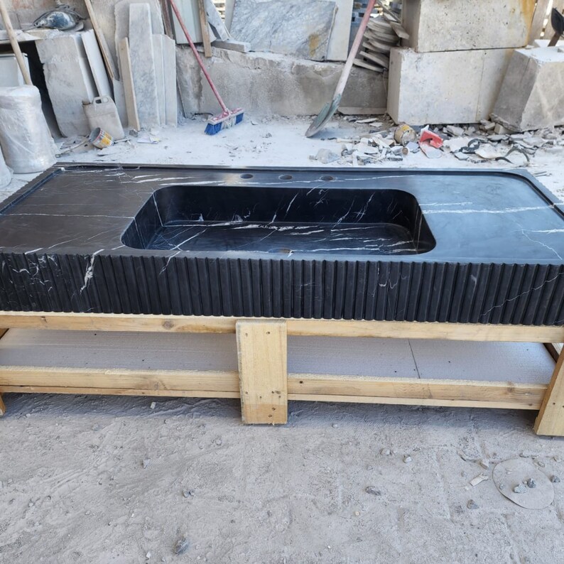 May include: A large, rectangular black marble countertop with a built-in sink basin. The countertop features white veining and a grooved front edge. It rests on a wooden pallet in an outdoor setting.