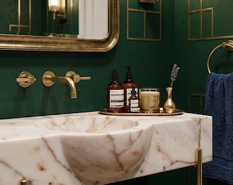 May include: A luxurious bathroom vanity with a marble sink and gold fixtures. A gold tray holds brown bottles, a candle, and a small vase with lavender. The mirror and wall sconces have gold frames, set against a dark green wall.