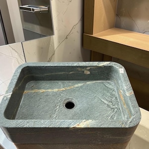 Peut inclure: Un &eacute;vier rectangulaire en pierre grise avec un trou de vidange central. L'&eacute;vier a une surface textur&eacute;e avec des stries dor&eacute;es et marron. L'&eacute;vier est plac&eacute; sur un fond neutre.