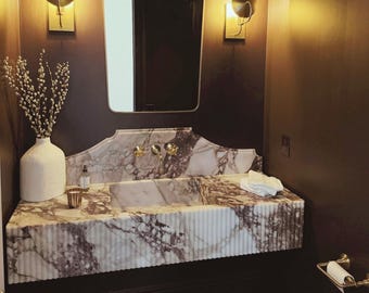 May include: A white marble bathroom vanity with a wavy edge, a white porcelain sink, and gold fixtures. The vanity is lit by two gold wall sconces with round shades. The walls are painted a dark brown colour.
