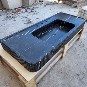 May include: A black marble bathroom vanity with a rectangular sink basin and three faucet holes. The vanity has white veining and a fluted edge design. It is resting on a wooden pallet.