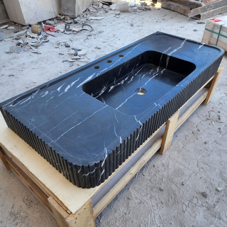 May include: A black marble bathroom vanity with a rectangular sink basin and three faucet holes. The vanity has white veining and a fluted edge design. It is resting on a wooden pallet.