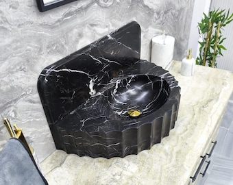 May include: A black marble sink with white veining, featuring a unique, angular design. The sink has a fluted base and a gold-colored drain. The bathroom fixture is set against a marble countertop and wall.