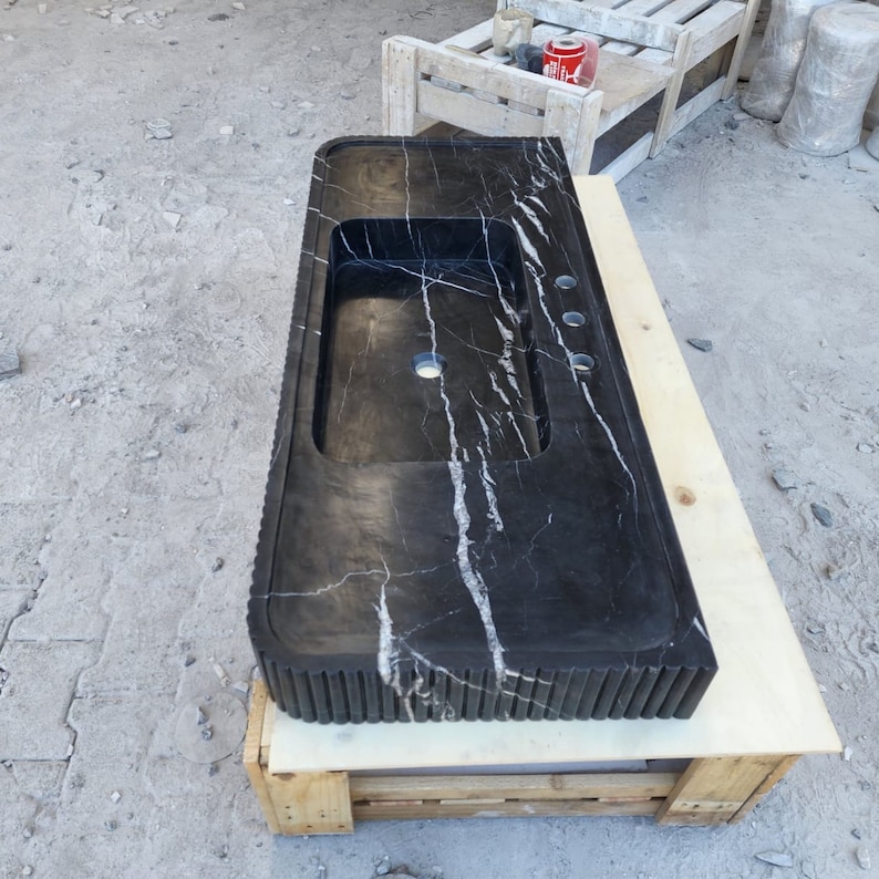 May include: A rectangular black marble countertop with a built-in sink and faucet holes. The countertop has white veining and a textured edge. It is resting on a wooden pallet.