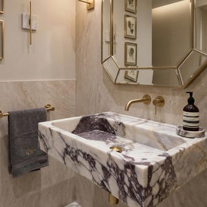 May include: A modern bathroom sink made of white marble with dark purple veining. The sink has a gold faucet and drain. A gold-framed mirror and a gray towel are also visible.