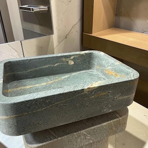 Peut inclure: Un &eacute;vier rectangulaire en pierre aux coins arrondis. L'&eacute;vier est de couleur gris-vert mouchet&eacute; avec des stries de beige et d'or. L'&eacute;vier est pos&eacute; sur une surface blanche. L'&eacute;vier est de conception moderne.