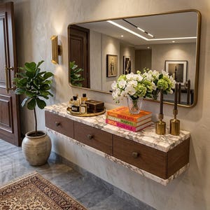 May include: A floating wall-mounted console table with a white marble top and three dark wood drawers. It features a large rectangular gold-framed mirror, a glass vase with white orchids, stacked books, gold candle holders, and a gold tray.