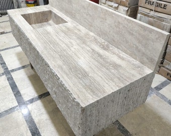 May include: A rectangular, beige stone bathroom vanity with a built-in sink. The vanity has a textured, rough-hewn exterior and a smooth, polished top. A backsplash is attached to the back of the vanity. The sink is rectangular.