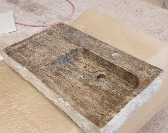 May include: A rectangular stone bathroom sink with a built-in basin and a faucet hole. The sink has a natural, textured appearance with shades of brown, tan, and gray. The edges are rough-hewn, giving it a rustic look.
