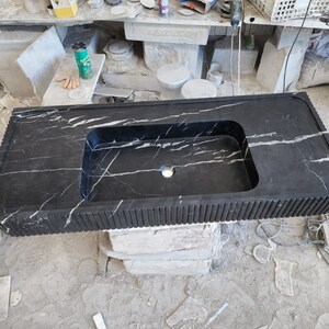 May include: A black marble countertop with a built-in rectangular sink. The countertop has white veining and a ribbed edge design. The sink has a single faucet hole. The countertop is resting on a stone base.